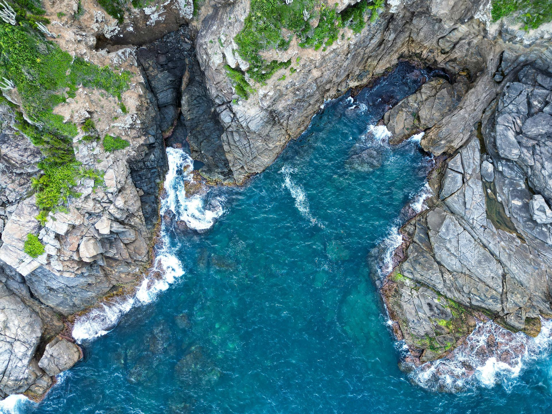 Vista aérea de um litoral rochoso com água turquesa. As ondas quebram na costa e a vegetação verde cresce nas falésias.