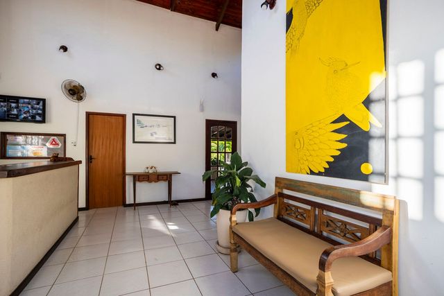 Hall de entrada com balcão de recepção, banco e obras de arte. Pintura abstrata amarela e preta na parede da direita. Piso de azulejo branco.