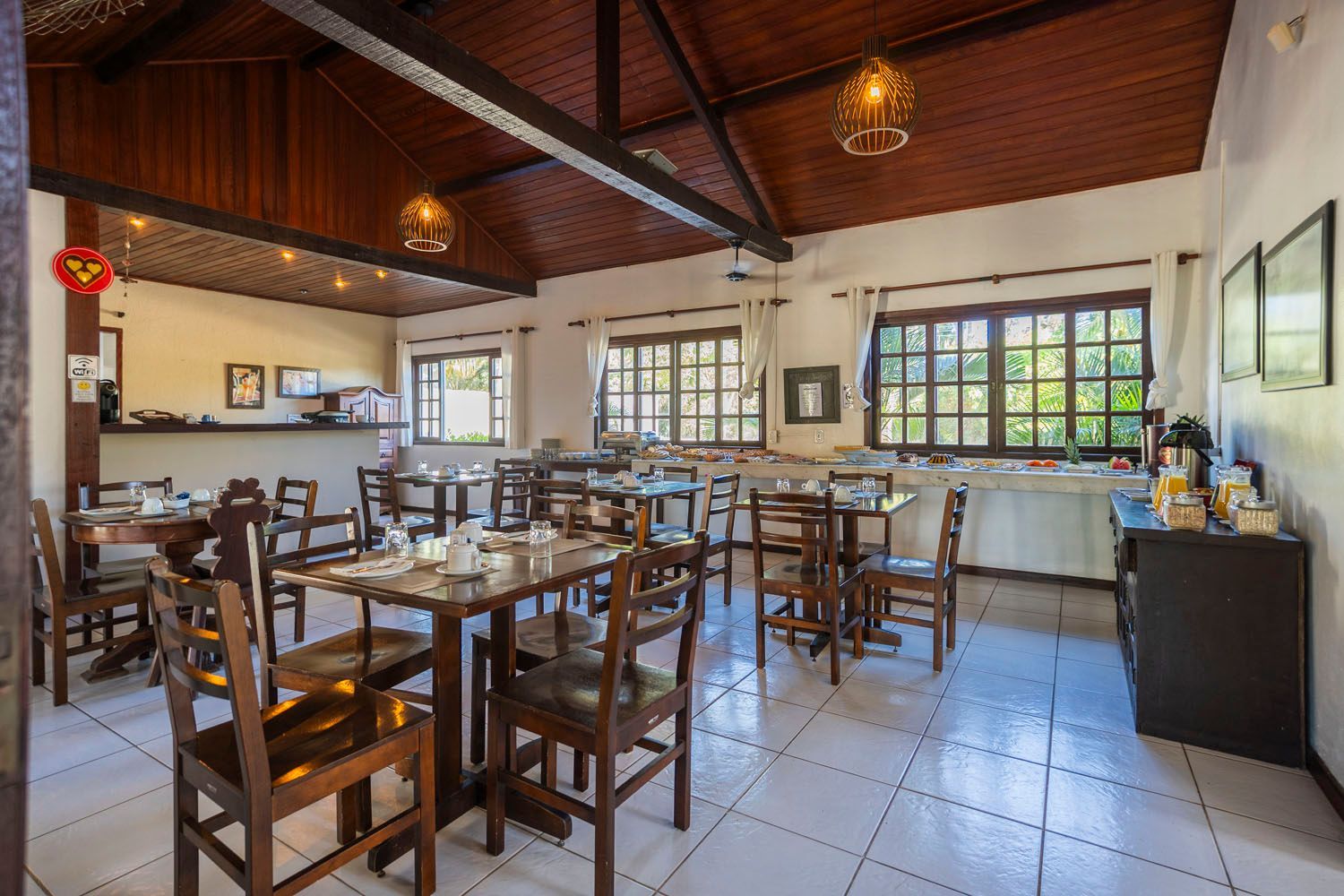 Sala de jantar com mesas e cadeiras de madeira, piso de azulejos brancos, teto e molduras de madeira escura e janelas.