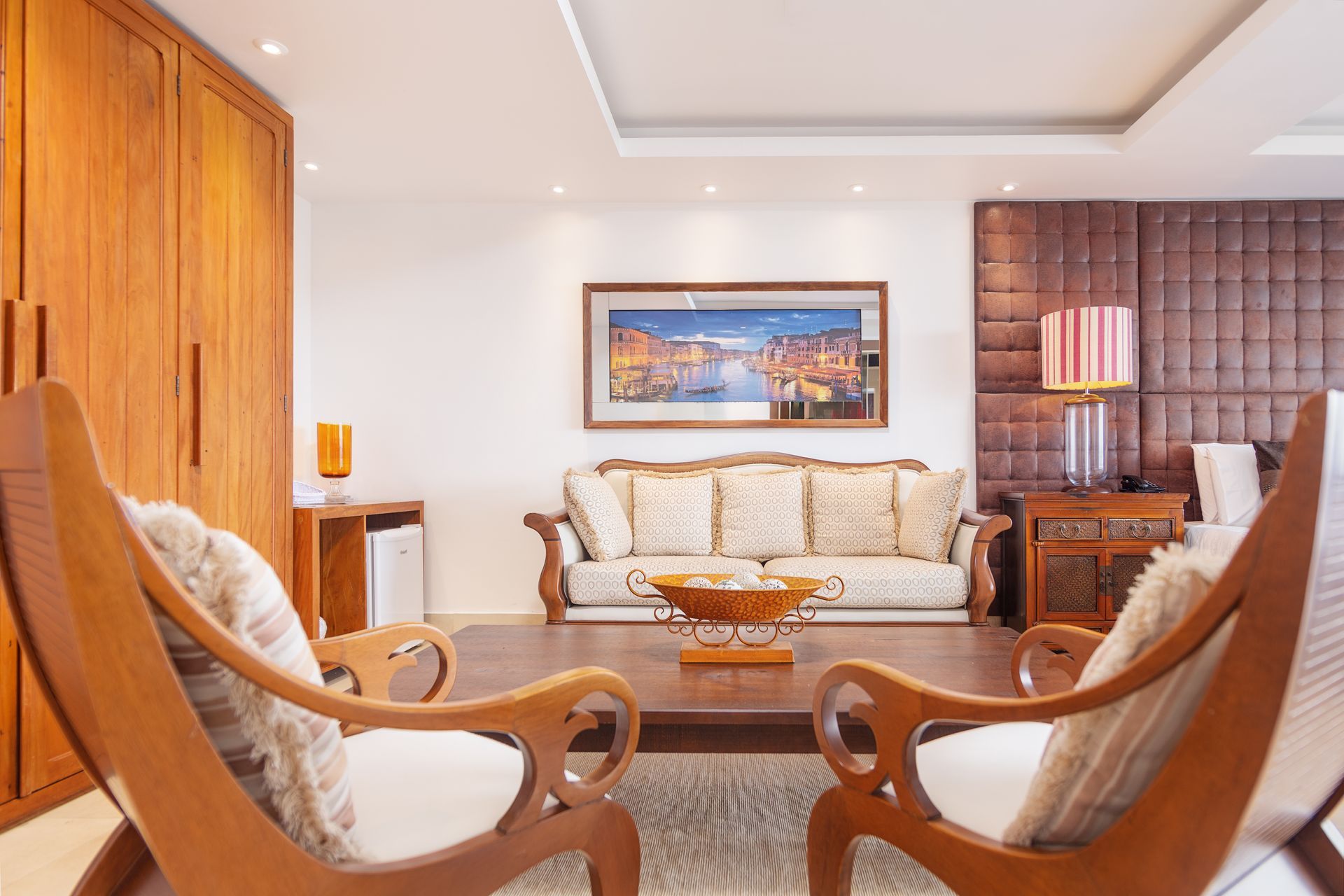 Sala de estar com móveis de madeira, sofá creme, obras de arte e duas cadeiras de madeira.