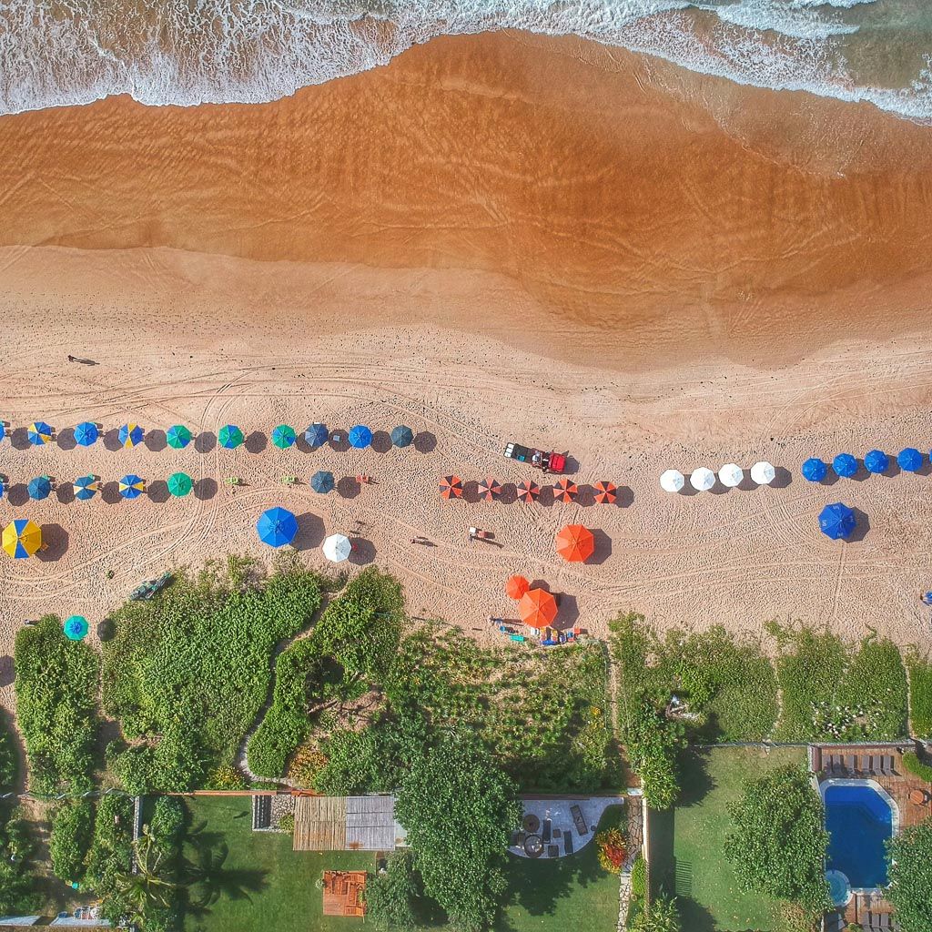 Vista aérea de uma praia com areia, oceano, guarda-sóis coloridos e vegetação verde com uma piscina.