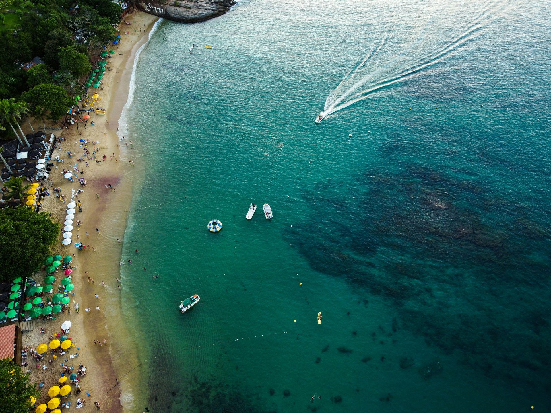 Vista aérea de uma praia com água turquesa. Barcos, pessoas e guarda-sóis verdes pontilham a orla. Uma lancha deixa um rastro na água.