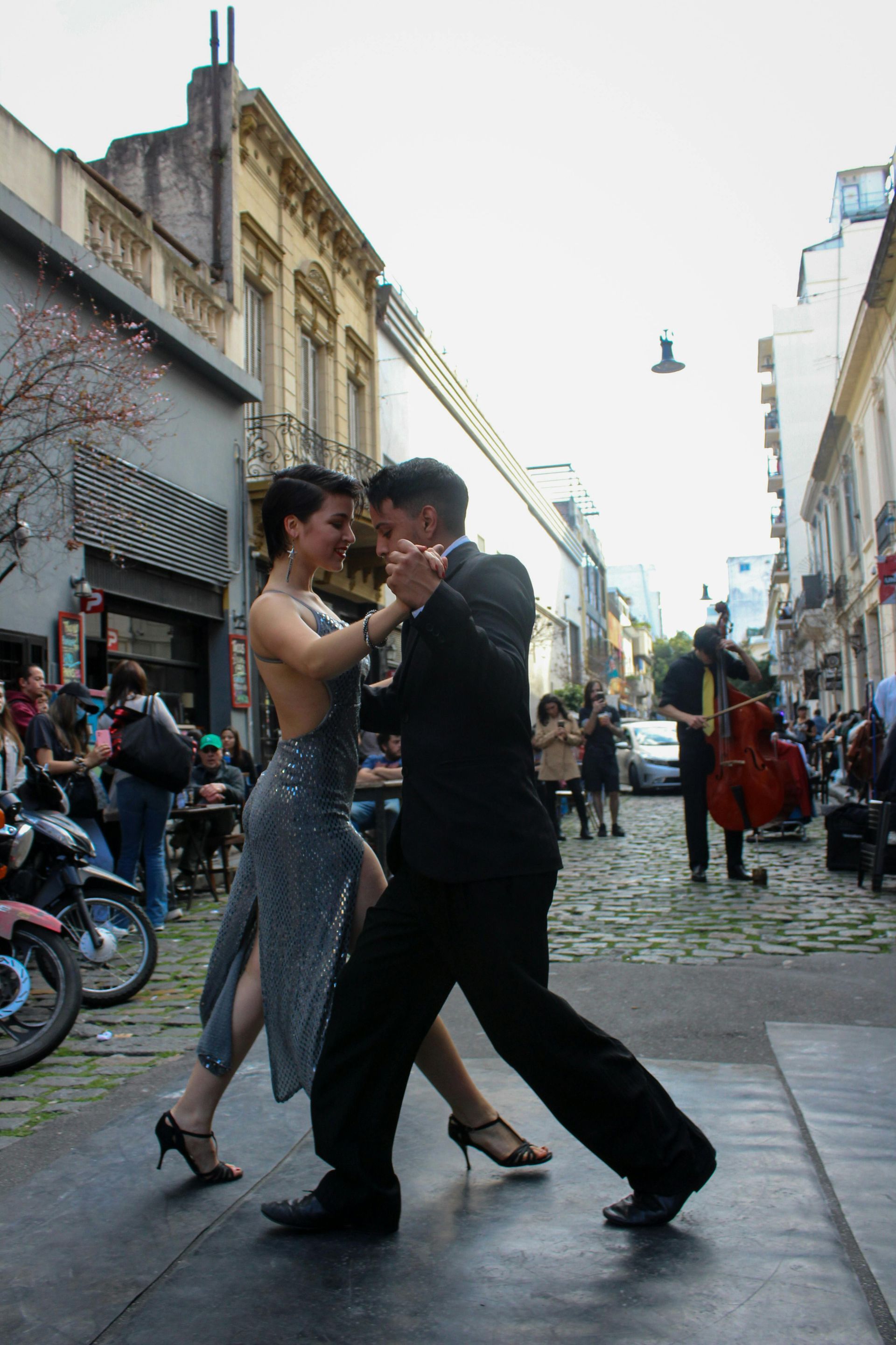 Two dancers in black and silver tango on a cobblestone street lined with spectators.