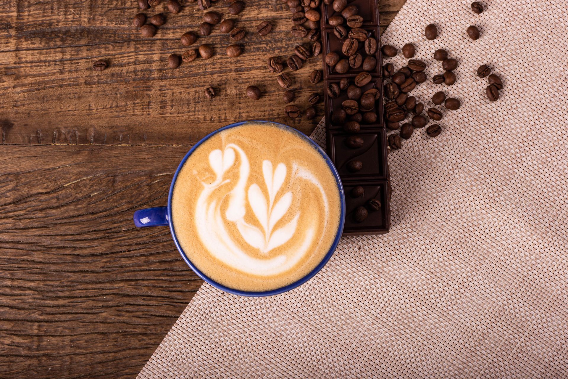 Xícara azul de latte com desenho de folha sobre uma mesa rústica de madeira, ao lado de grãos de café e um pano de juta.