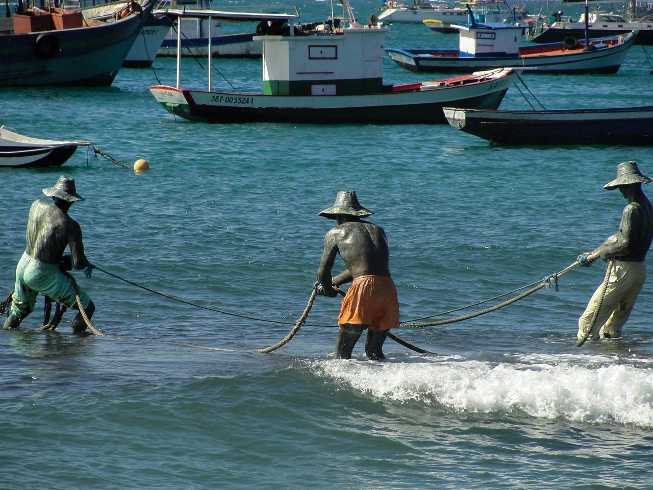 Estátuas de três figuras puxando uma rede do mar, com barcos ao fundo.