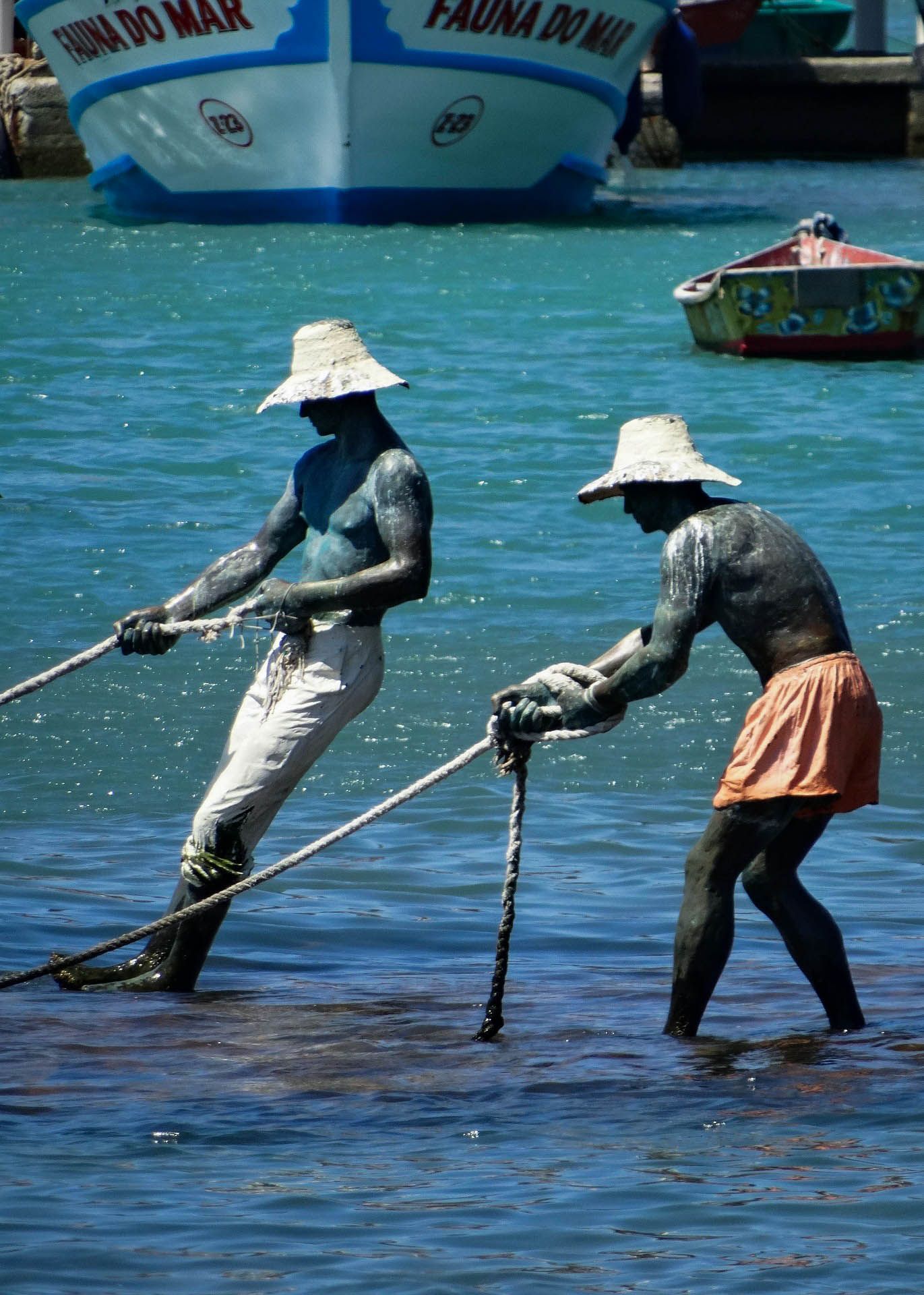 Duas pessoas puxam cordas da água perto de barcos. Elas usam chapéus e estão cobertas com tecido escuro.