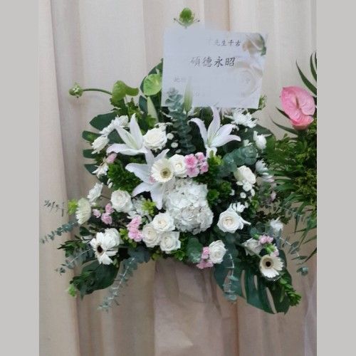 一束以白色百合、玫瑰和綠葉為主的花束，擺放在窗簾前。旁邊放著一張卡片。