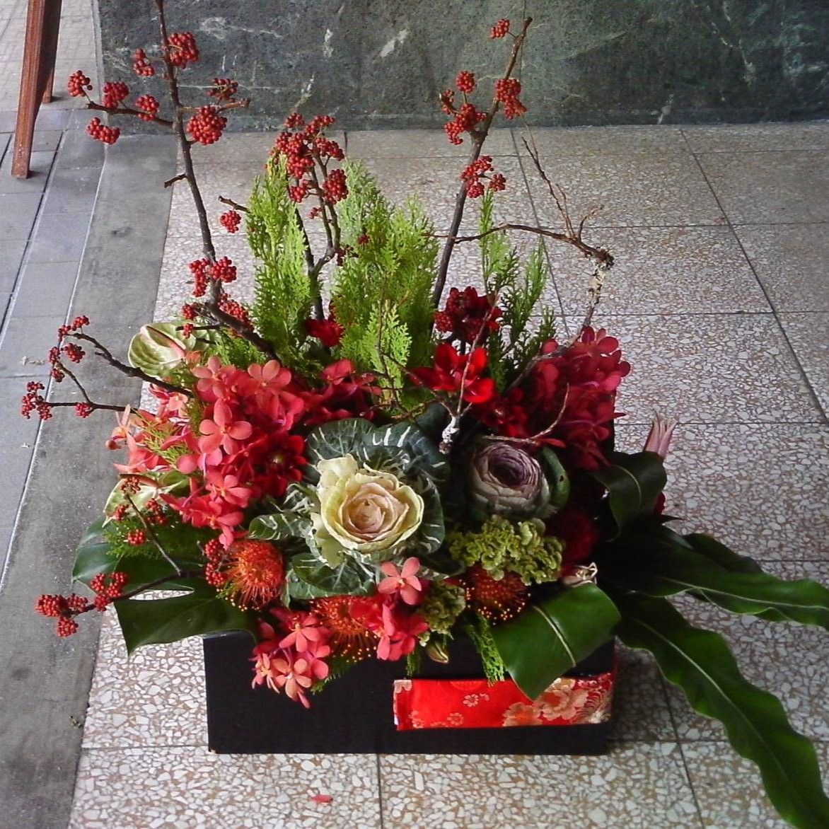 黑色長方形盒子裡擺放著一束花卉，花材為紅色、綠色和奶油色的花朵和葉子。