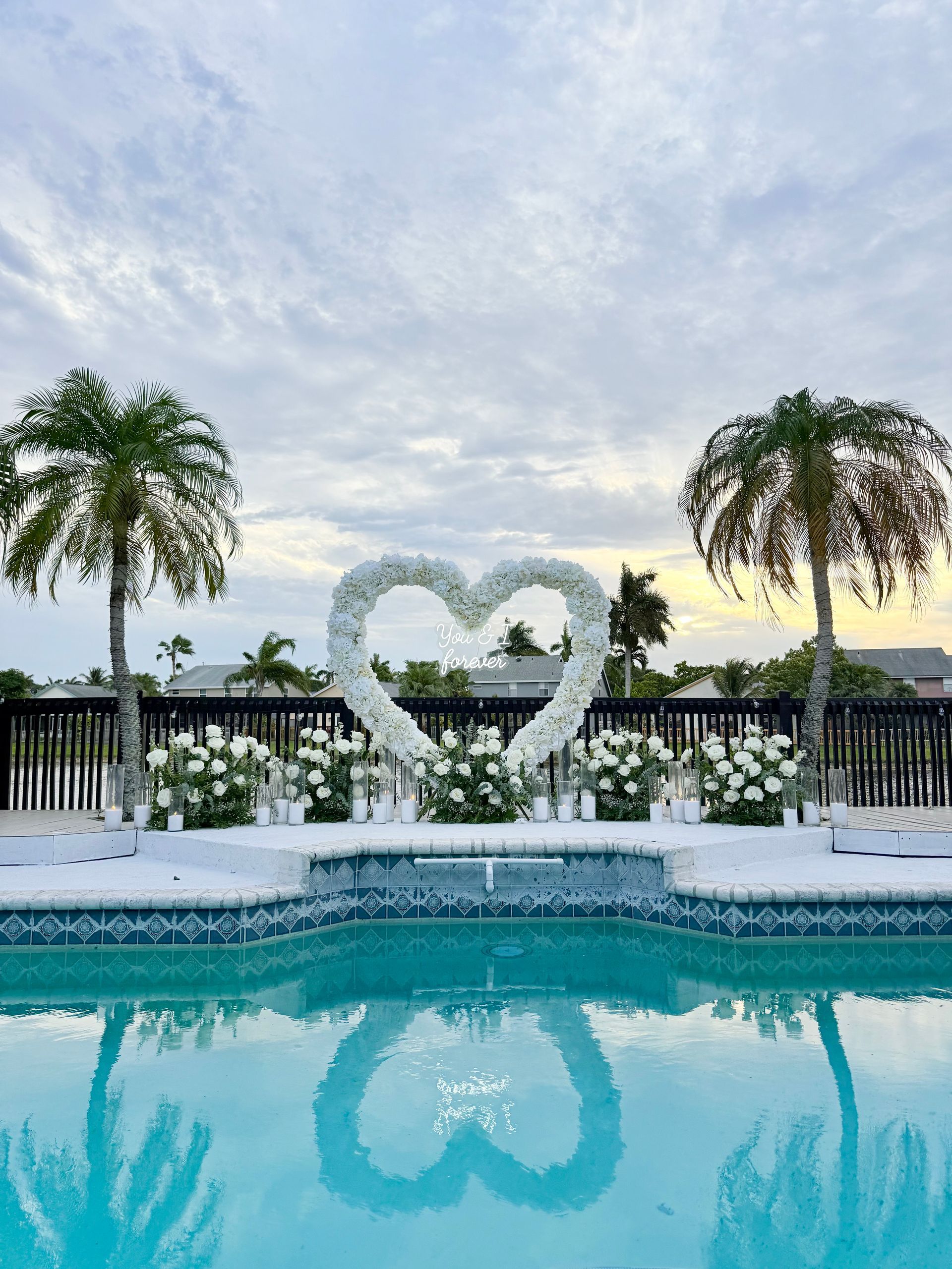 Heart-shaped floral arrangement by a pool; palm trees, cloudy sky reflected in water.