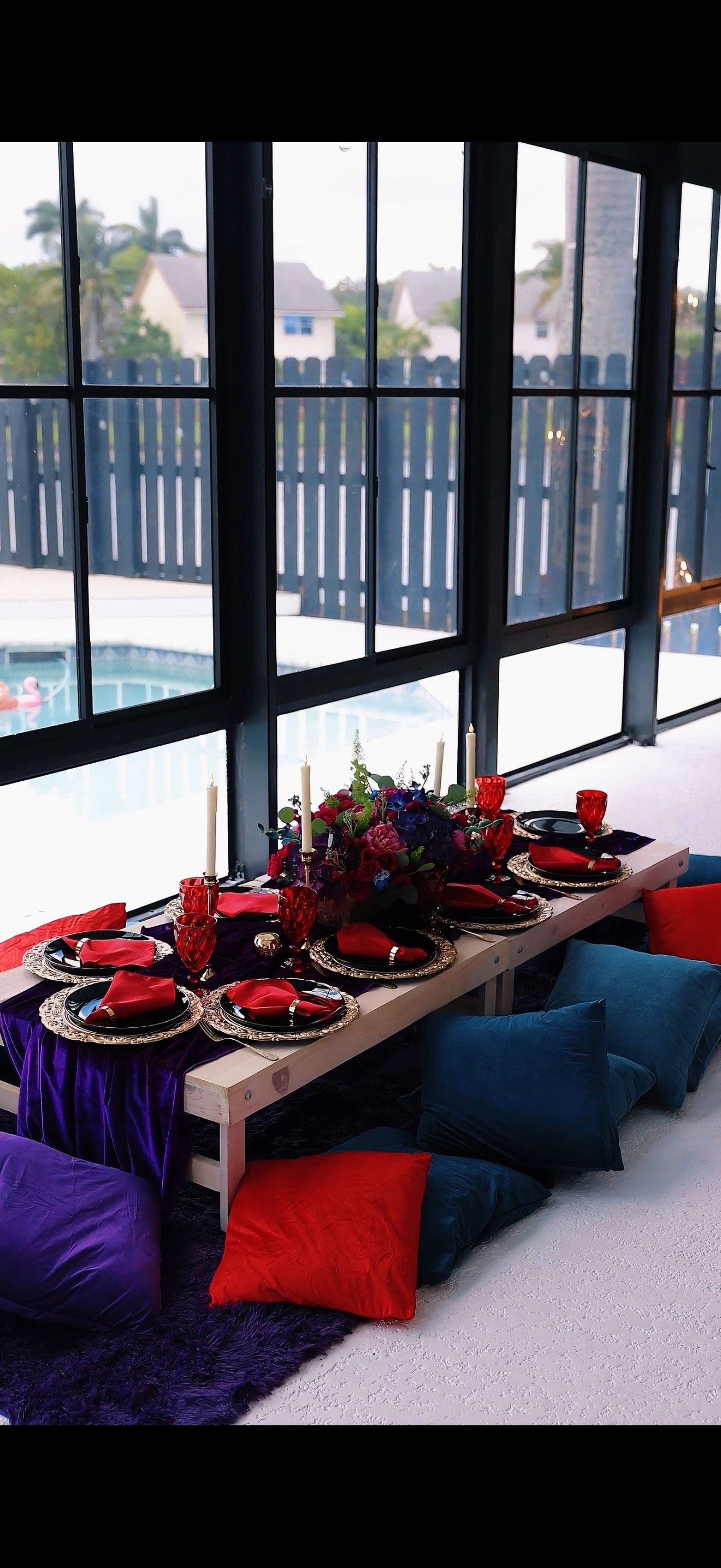 A low table set for dining, with red and purple decor. Pillows on the floor, view of pool outside.