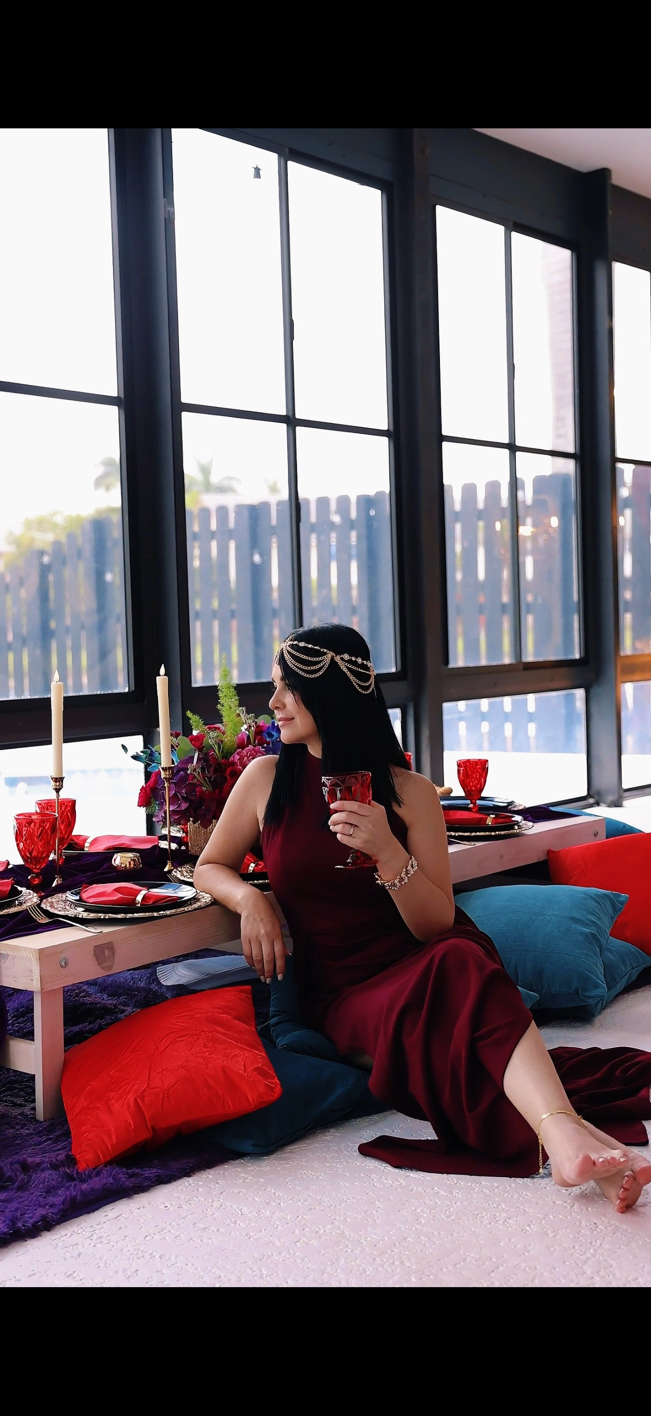 Woman in a burgundy dress sits on cushions next to a low table with red decor. Large windows in background.