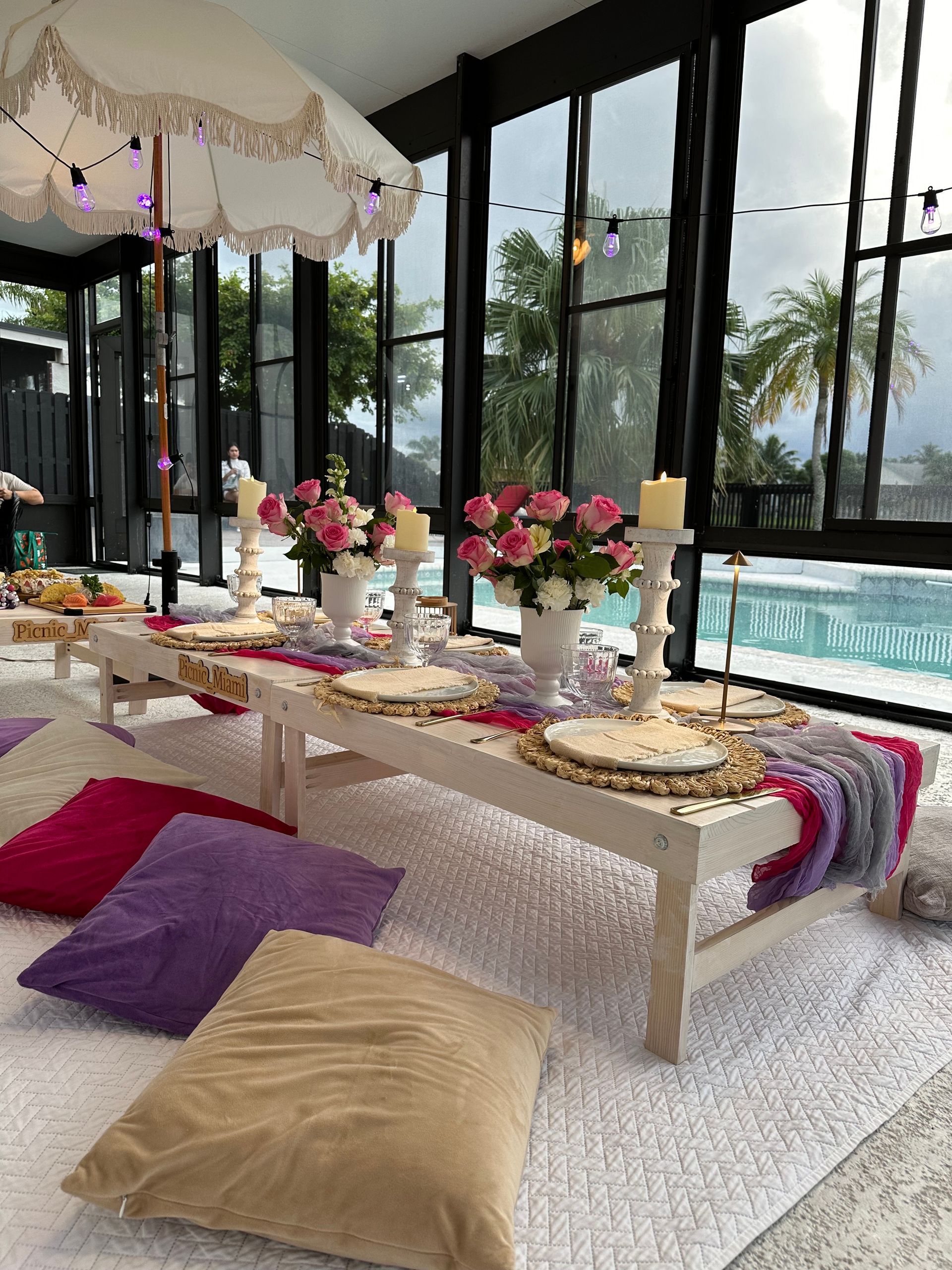 Low picnic table set for a party with flowers, candles, cushions, and an umbrella, next to a pool.