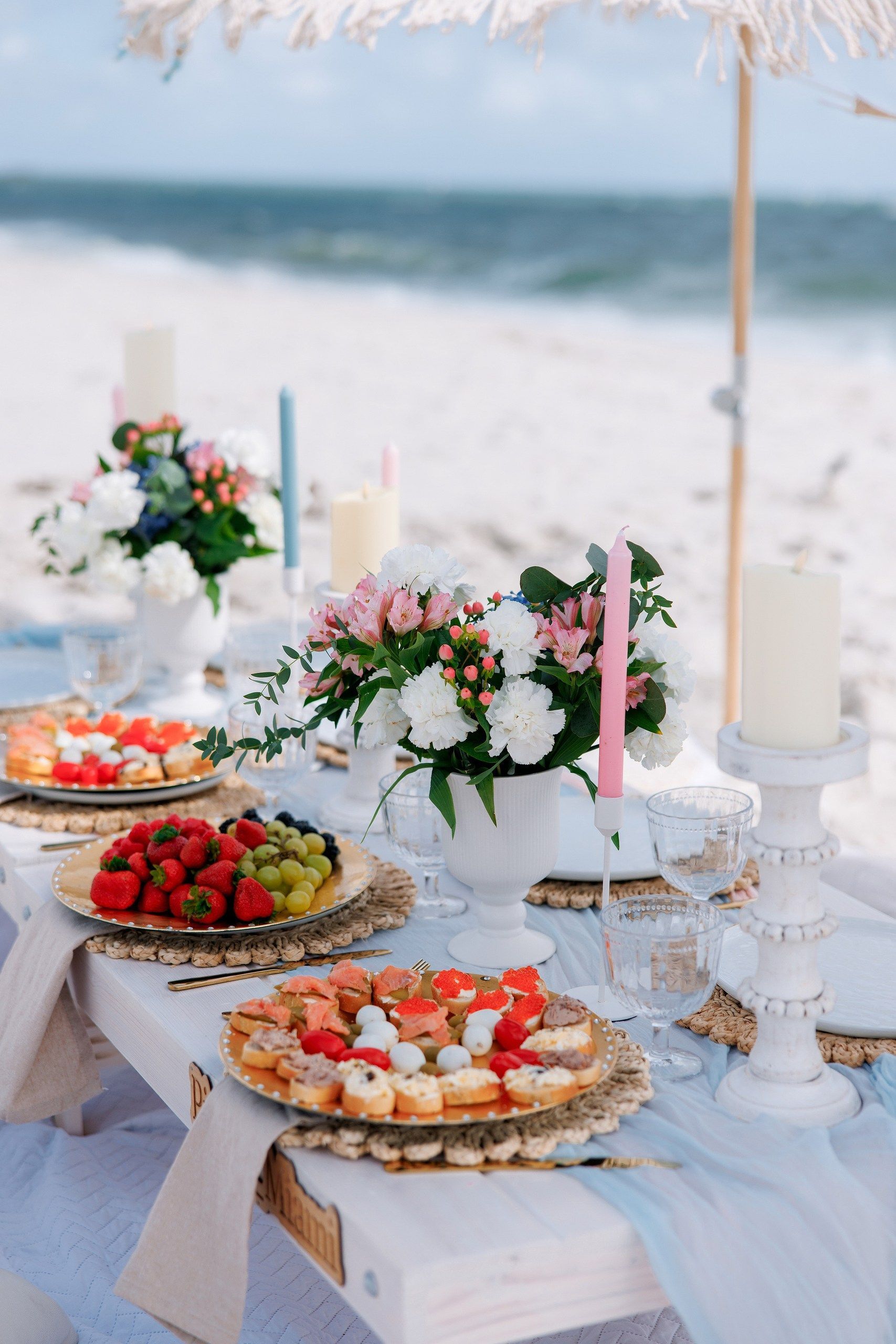 Beach picnic setup with flowers, fruit plates, candles, and a blue runner on a low white table.