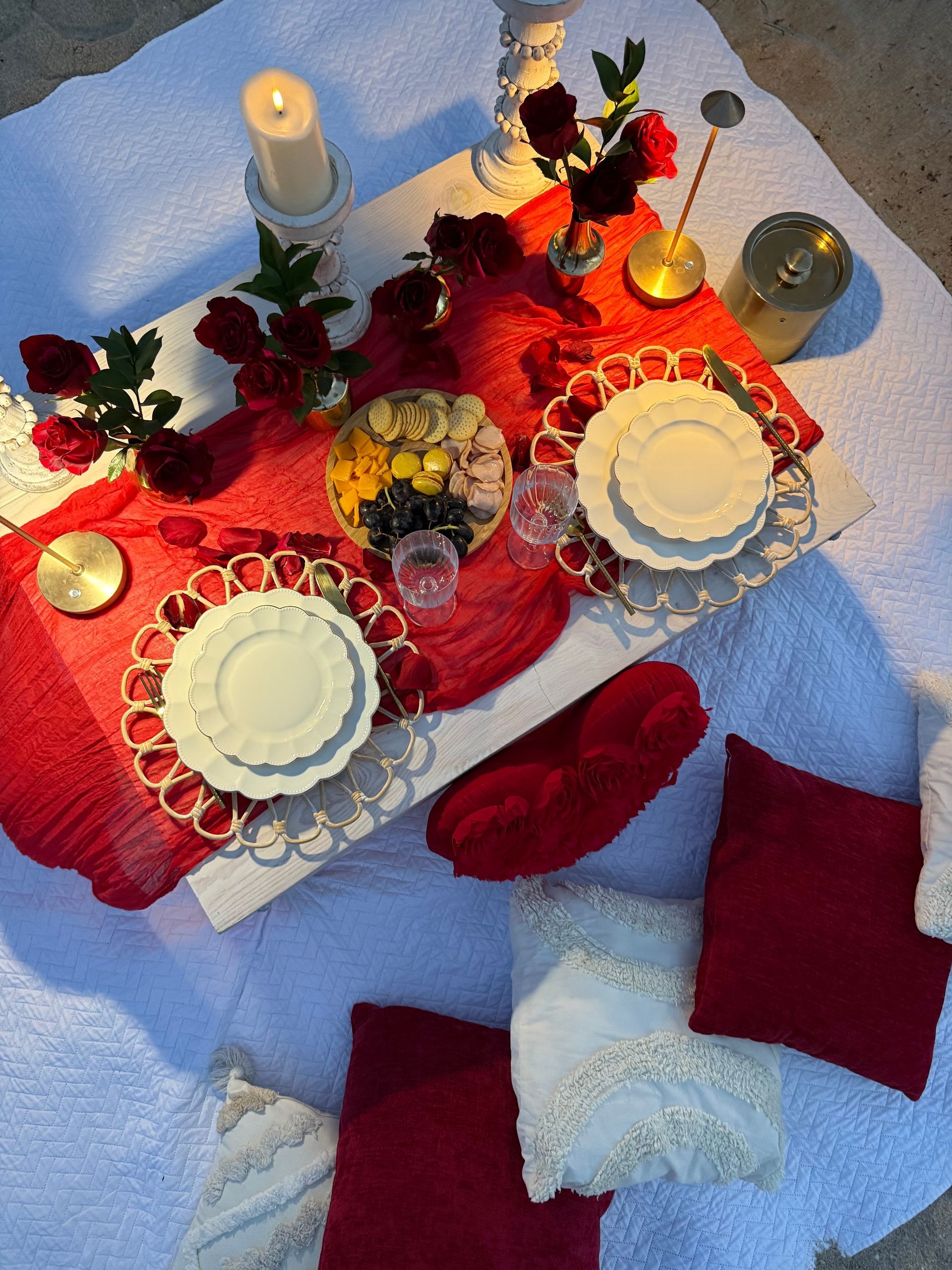 Romantic beach picnic setting: red runner, floral centerpieces, white plates, candles, and pillows.