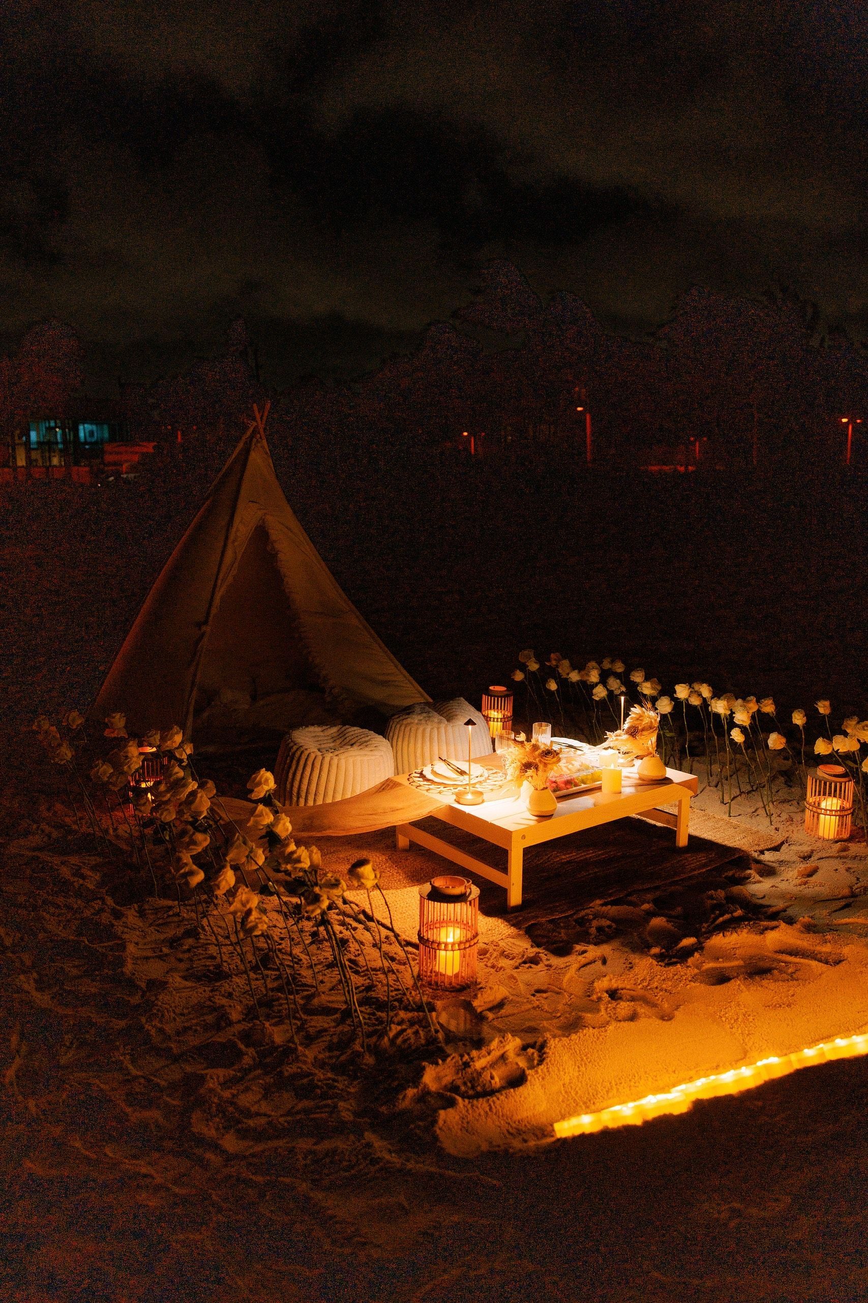 Romantic beach dinner setup at night with lit lanterns and a tent.