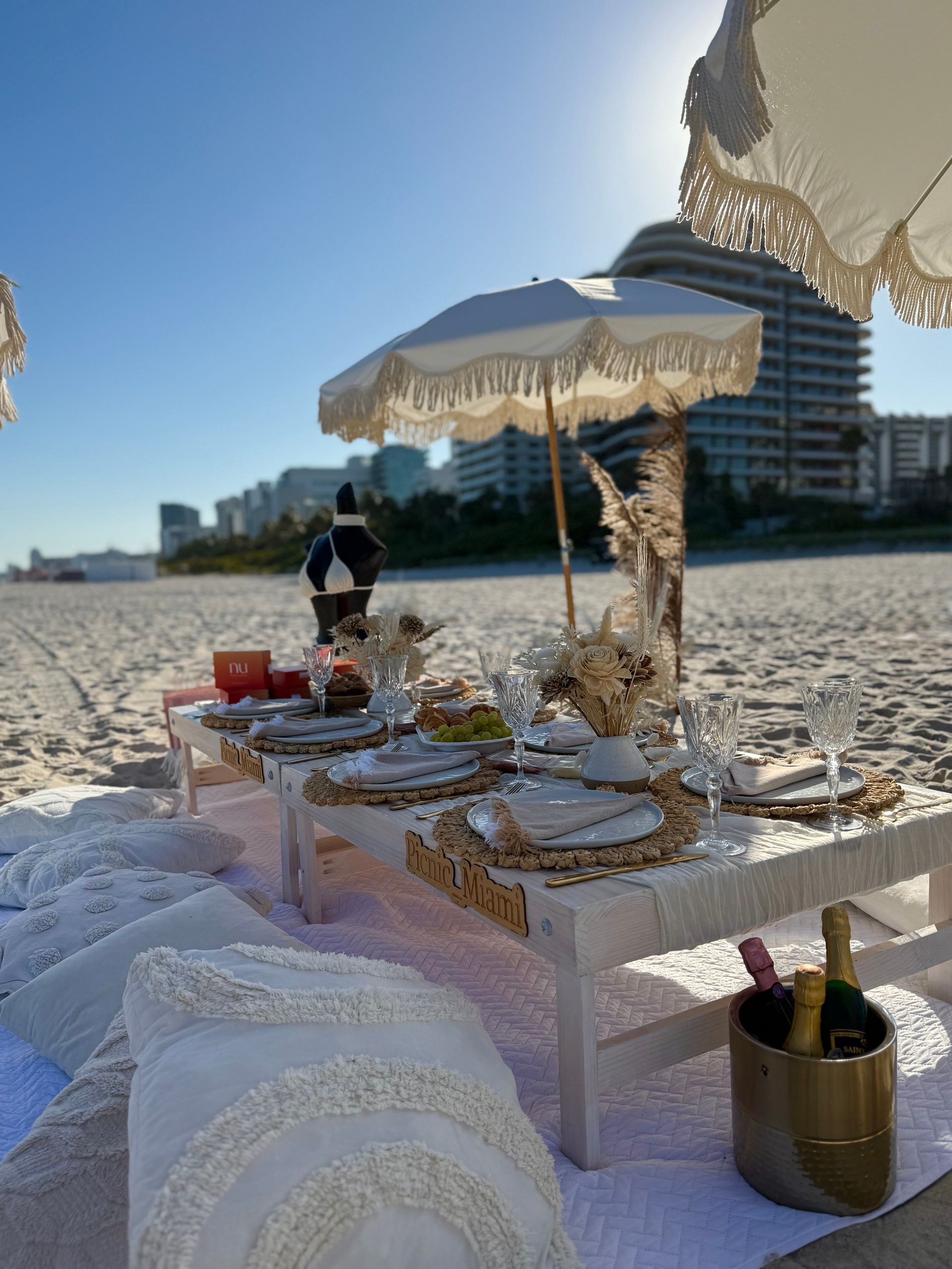 Beach picnic setup with white table, decor, pillows, and sun umbrellas.