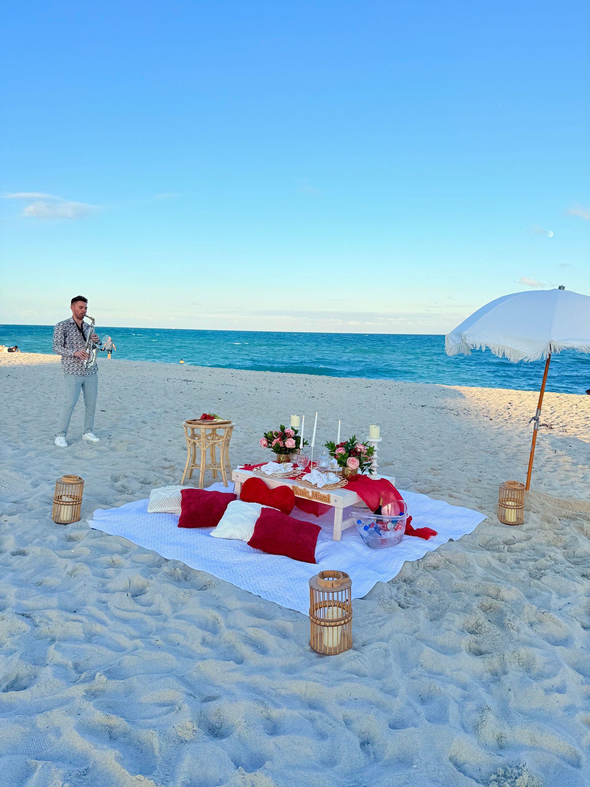 Beach picnic set up with food, flowers, and a person holding a bouquet, near the ocean.