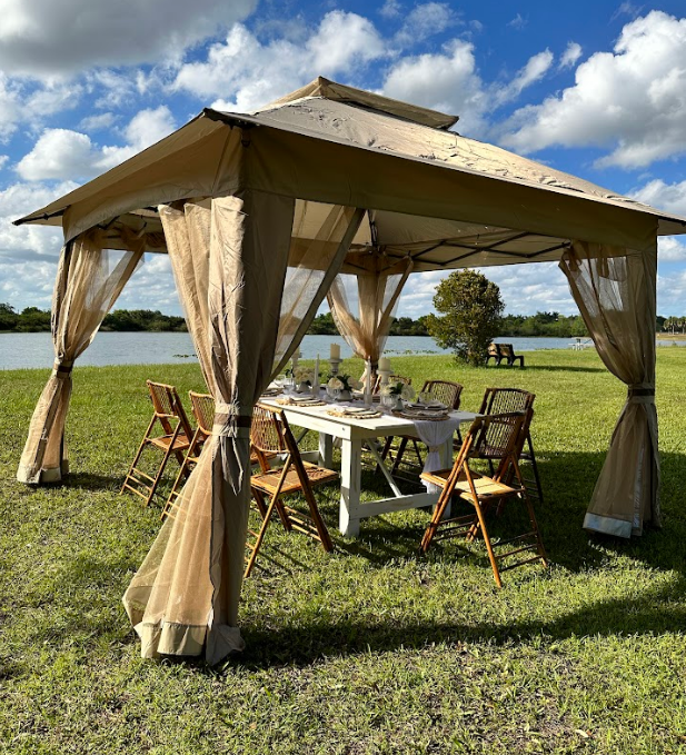 Gazebo with draped curtains over a white table set for a meal, chairs surrounding it, on grass near a lake.