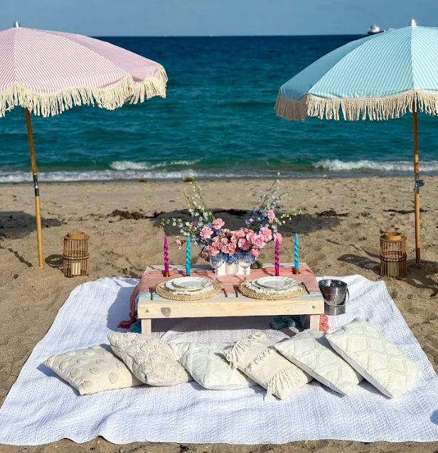 Beach picnic with pink and blue umbrellas, table set with flowers, pillows, and the ocean in the background.