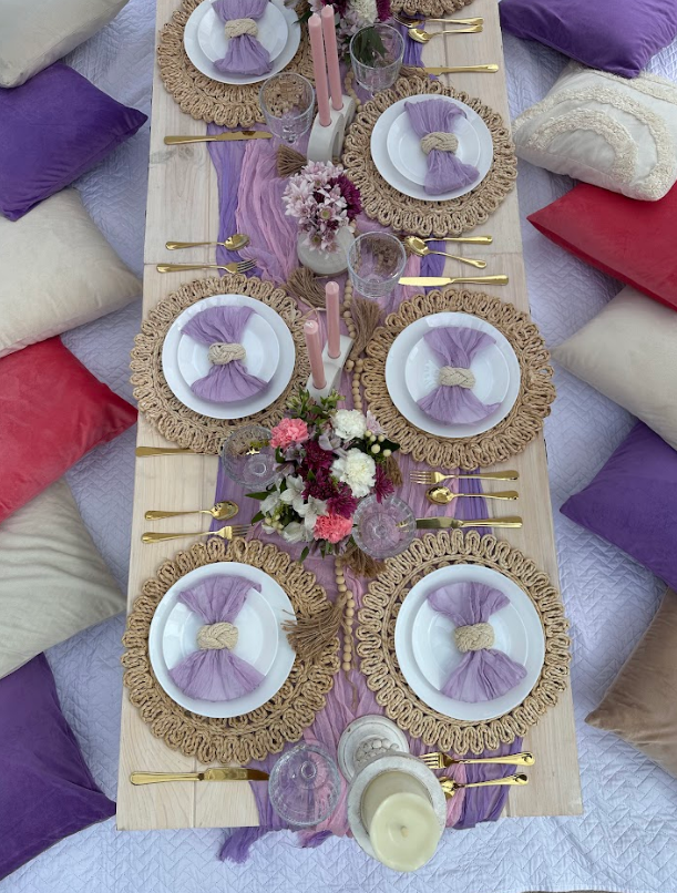 Picnic table set with purple and gold tableware, surrounded by pillows.