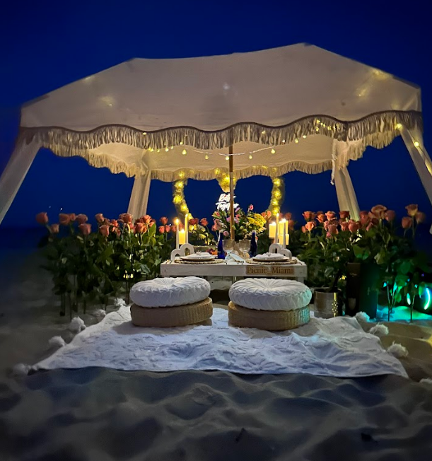Romantic beach dinner setting with canopy, roses, and candles at night.