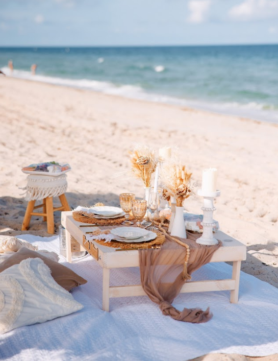 Beach picnic setup with low table, plates, candles, and decorative elements on a sandy beach with ocean view.