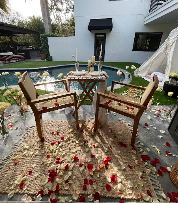 Romantic outdoor setting: table, chairs, rug, rose petals near pool and white tent.