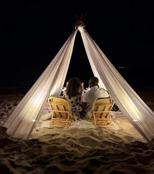 Couple seated under a draped fabric structure on a beach at night; lit by candles, with chairs.