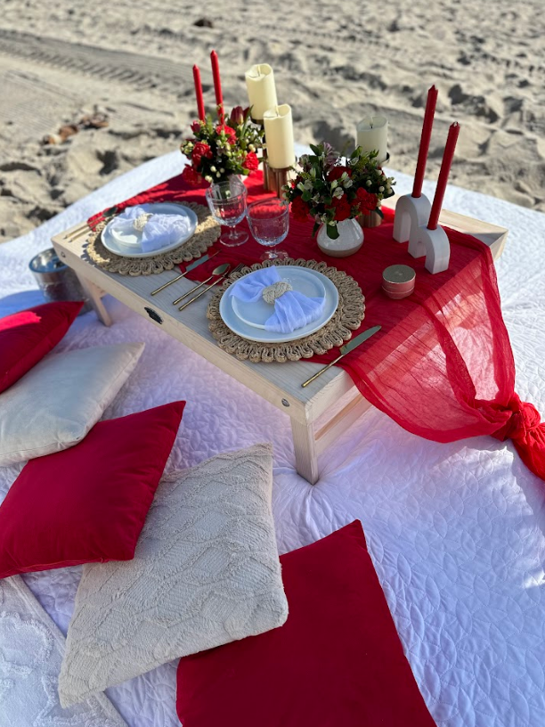 Beach picnic setup with red and cream decor: table, pillows, candles, flowers, plates, and glasses.