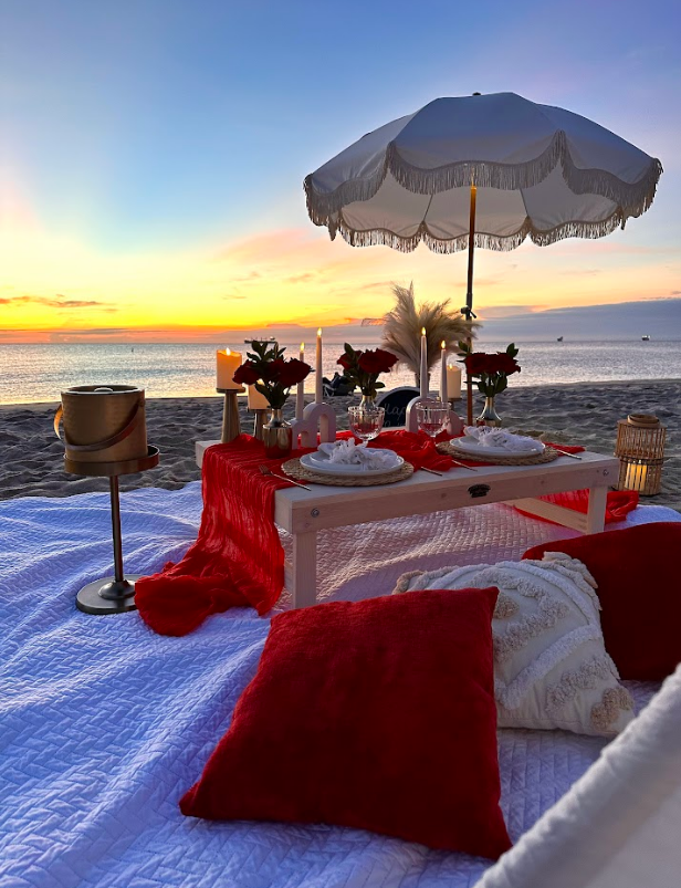 Romantic beach picnic setup with red and white decor; sunset over ocean.