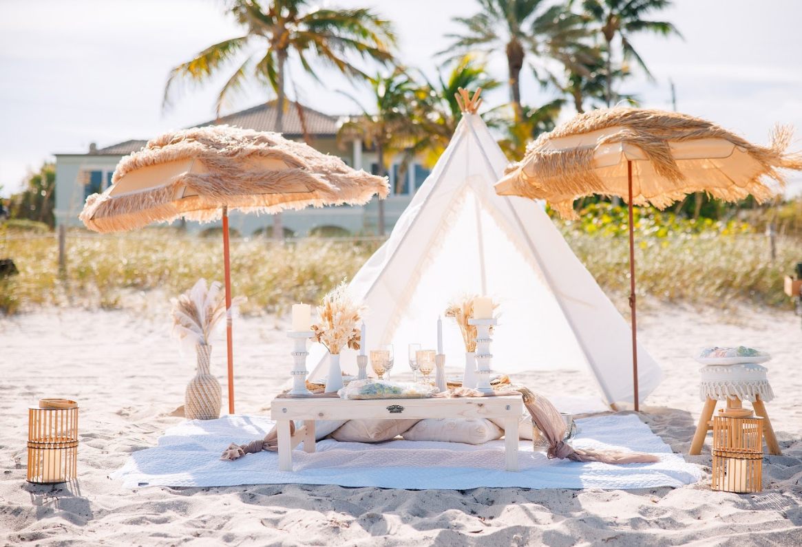 Beach picnic setting with fringed umbrellas, teepee, and table with decor.