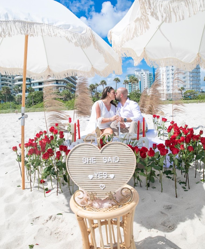 Beach proposal: couple kissing under umbrellas, red rose path, heart sign says