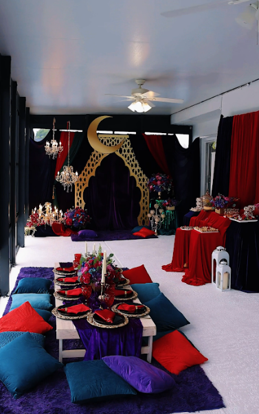 An Arabian Nights-themed party with low seating, red, purple, and gold decor, and a large gold crescent moon backdrop.
