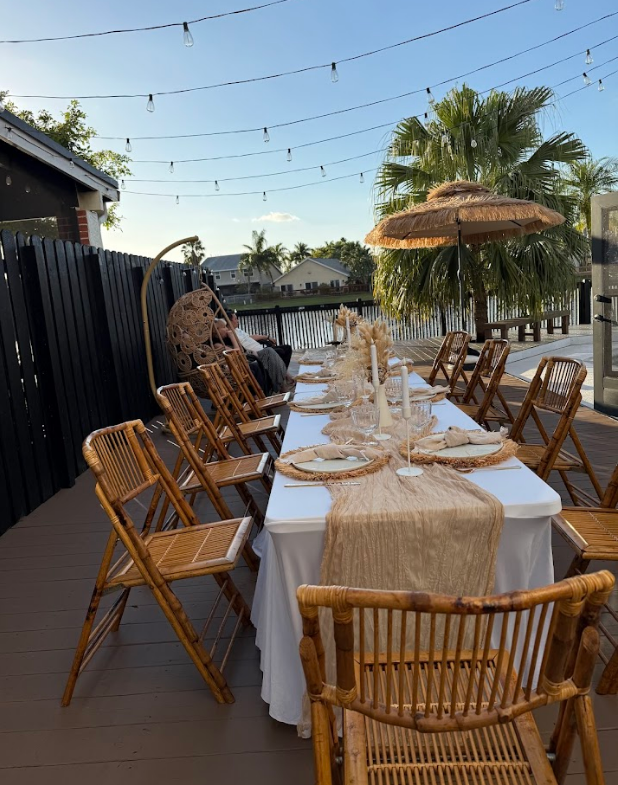 Outdoor dining table set for a gathering; wooden chairs, white tablecloth, string lights, and a thatched umbrella.