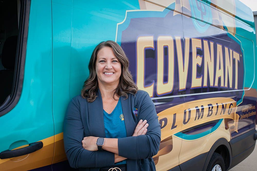 Smiling woman in blue jacket standing beside a turquoise Covenant Plumbing van logo