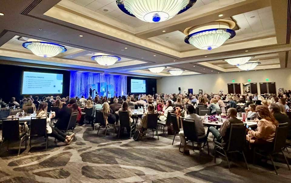 Conference banquet with attendees seated at round tables, blue-lit stage, and large presentation screens