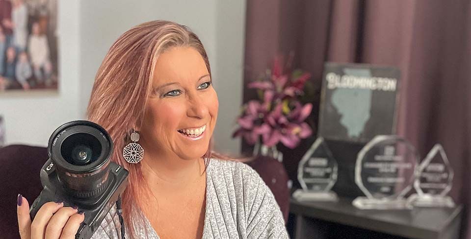 Woman holding a camera in a bright office, smiling beside awards on a shelf.