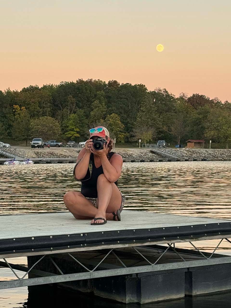 Person kneeling on a dock, taking a photo by a calm lake at sunset with trees and the moon in the background.