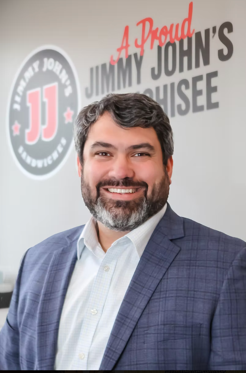 Smiling man in a blue suit standing in front of a Jimmy John’s Proud House sign.