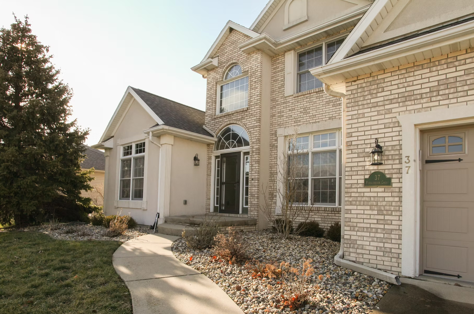 Suburban house exterior with beige brick, front entry, windows, and a small landscaped walkway