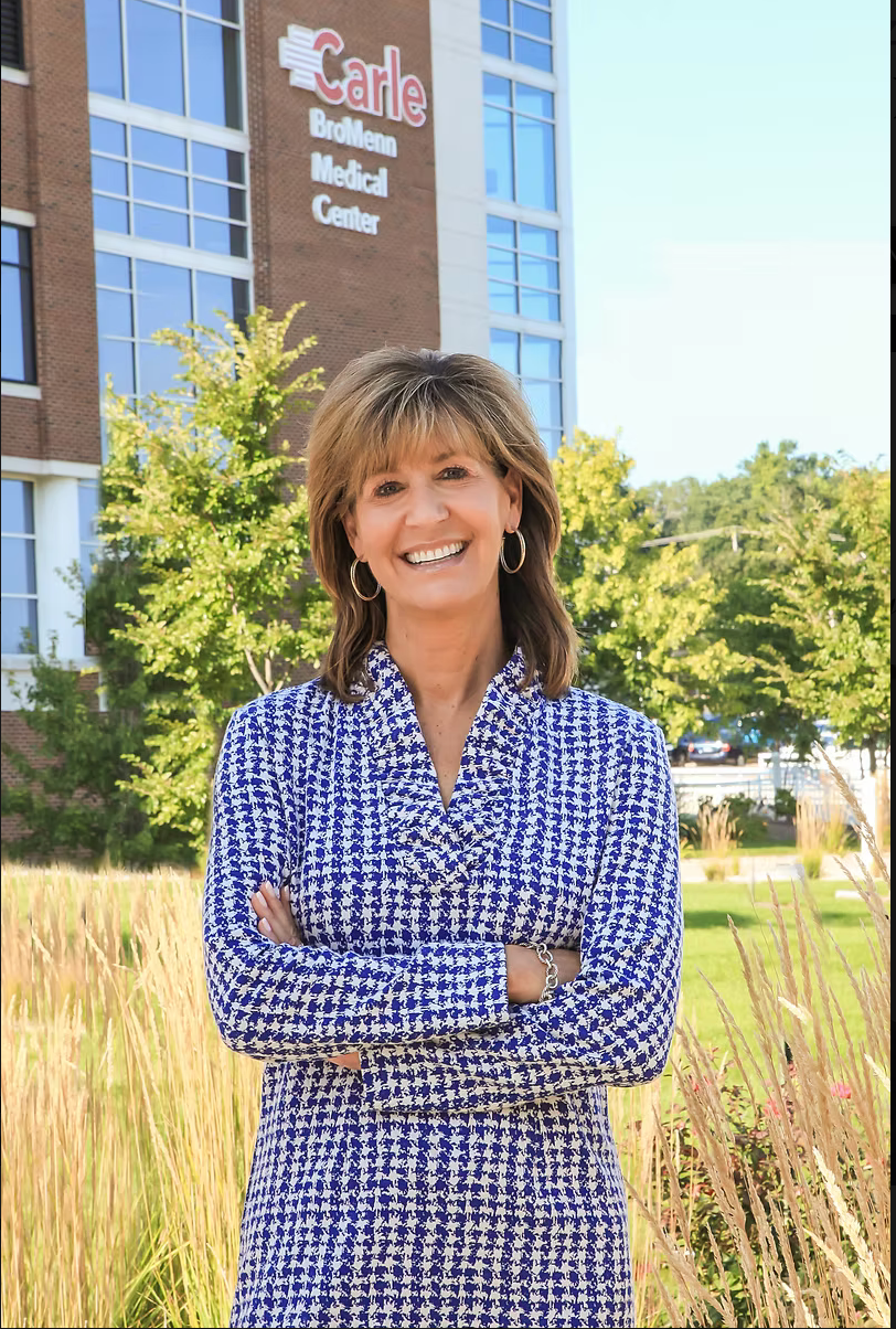 Smiling woman in blue patterned dress stands with arms crossed outside a building with tall grasses.
