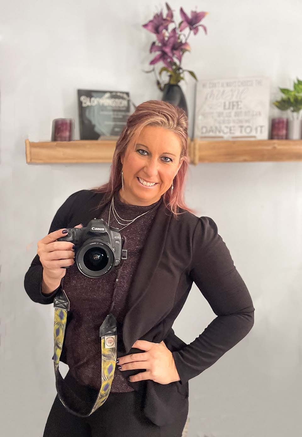 Woman smiling, holding a camera beside a floating shelf with decor and flowers in a bright room