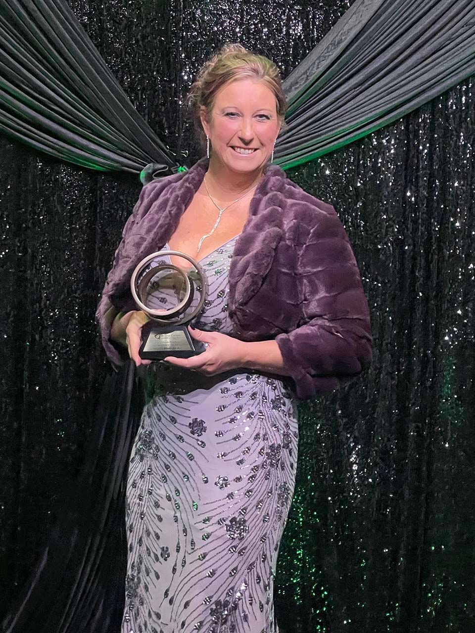 Woman holding a trophy, wearing a purple jacket and floral dress, standing before a dark draped backdrop.