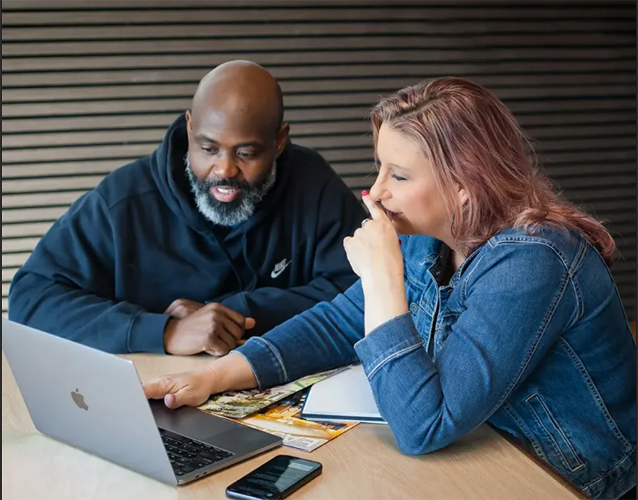 Two coworkers review notes on a laptop at a table in a meeting room