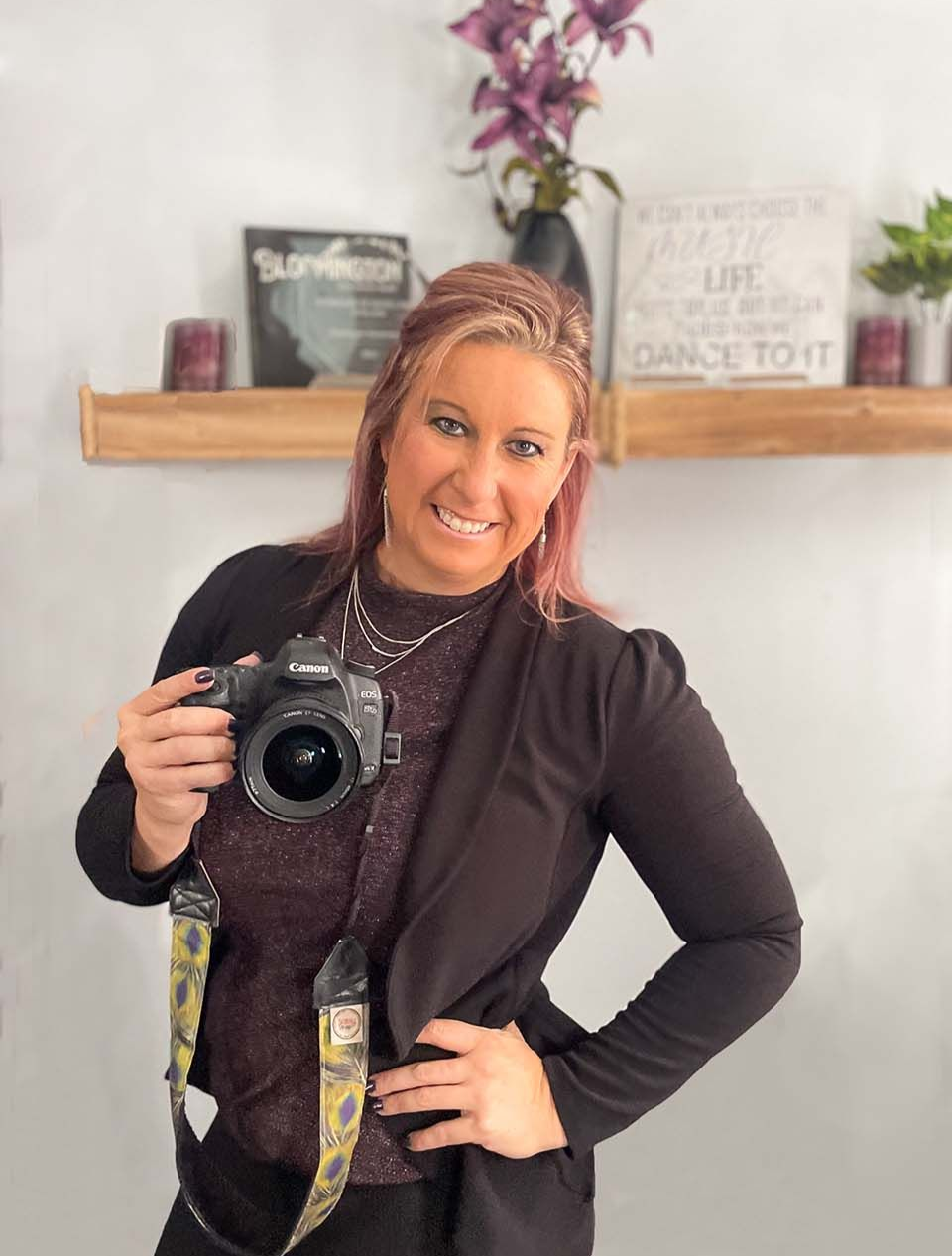 Smiling woman holding a camera in a bright room with a shelf and flowers behind her