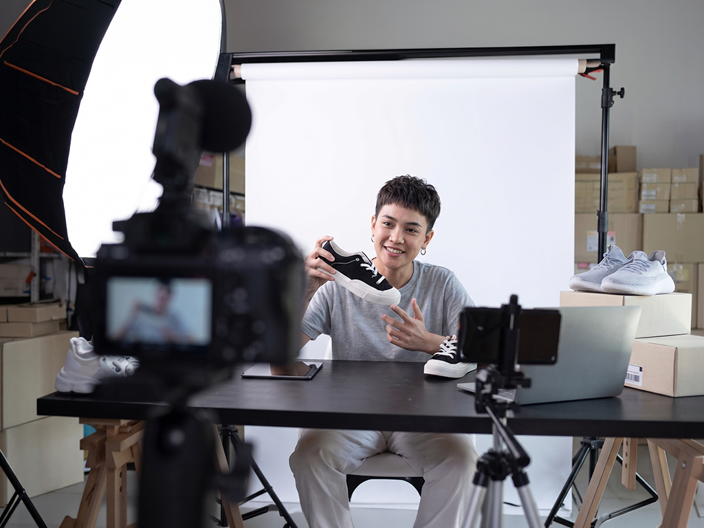 Person filming a smiling speaker at a table in a studio with a white backdrop and camera equipment.