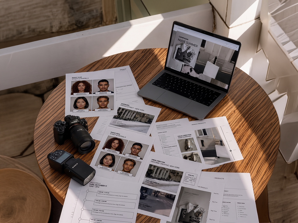 Laptop, camera gear, and printed photo proofs spread across a wooden table in a bright room