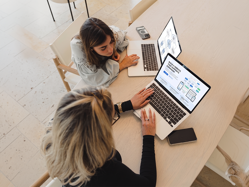 Two people collaborate at a table with laptops in a bright room.