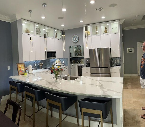 Modern kitchen with white cabinets, marble countertop island, and blue bar stools. Person in frame.