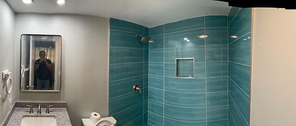 Bathroom with teal tiled shower, mirror, and vanity. A person is reflected in the mirror.
